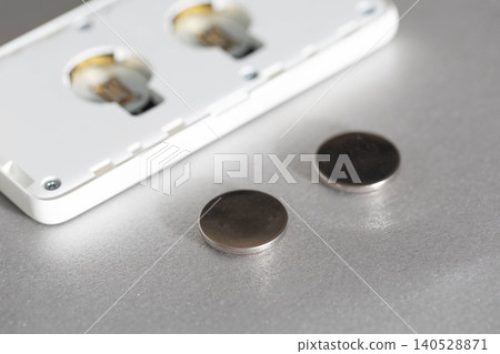 Button cell batteries lie on metallic surface, ready for replacement in open white electronic device, revealing empty compartments for power source maintenance Button cell batteries lie on metallic surface, ready for replacement in open white electronic device, revealing empty compartments for power source maintenance 140528871