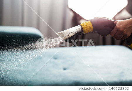 Person hand holding professional cleaning nozzle spraying water and solution onto fabric surface of blue sofa during deep upholstery washing and furniture care Person hand holding professional cleaning nozzle spraying water and solution onto fabric surface of blue sofa during deep upholstery washing and furniture care 140528892