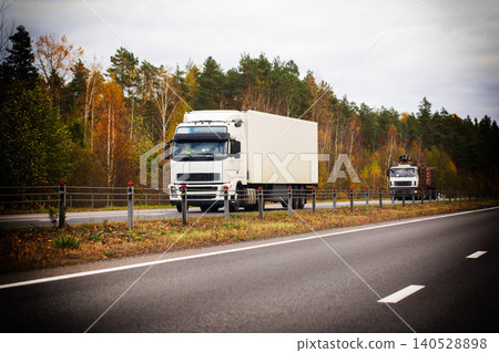 White semi-truck with cargo container drives on highway road during autumn season, logging truck follows, forest trees on roadside with fall foliage color White semi-truck with cargo container drives on highway road during autumn season, logging truck follows, forest trees on roadside with fall foliage color 140528898