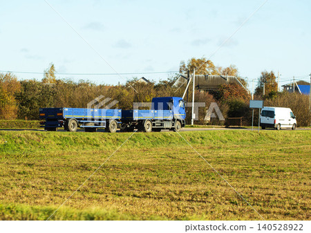 Blue truck and trailer driving on road beside grassy field and autumn foliage with white delivery van parked at roadside under clear sky during day Blue truck and trailer driving on road beside grassy field and autumn foliage with white delivery van parked at roadside under clear sky during day 140528922