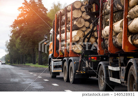 Large commercial logging truck transporting stacked cut wooden logs on an open asphalt country road lined with green trees during warm sunset light. Copy space for text Large commercial logging truck transporting stacked cut wooden logs on an open asphalt country road lined with green trees during warm sunset light. Copy space for text 140528931
