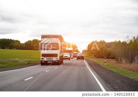Traffic flow of commercial trucks and passenger cars on asphalt road through rural landscape under overcast sky with sunlight flare 140528933