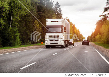 Large white semi truck driving on multi lane highway with other vehicles, surrounded by dense green forest trees, warm sunlight 140528934