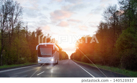 White passenger bus travels rural highway at golden hour, headlights illuminated, long journey road transportation service logistics infrastructure 140528937