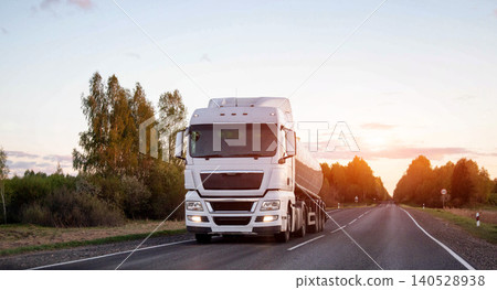 White modern semi truck with tanker trailer traveling on asphalt country road surrounded by trees during golden hour sunset light. 140528938