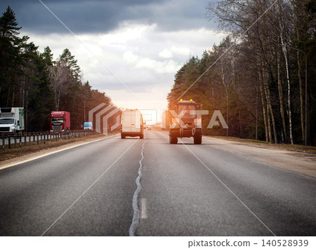 Vehicles on highway; tractor, cargo van, and semi-trucks traveling rural forest landscape under overcast sky, evening sunset. Copy space for text 140528939