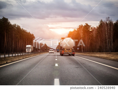 White tanker truck transporting flammable liquid on asphalt highway, surrounded by dense pine and birch forest, under overcast sky, showing essential cargo transportation, evening sunset. Copy space 140528940