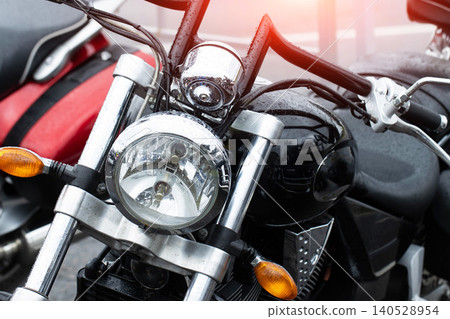 Close-up view of a wet black motorcycle front section with chrome headlight, handlebars, and signal lights, covered in raindrops after rain, showcasing cruiser style and robust engineering 140528954