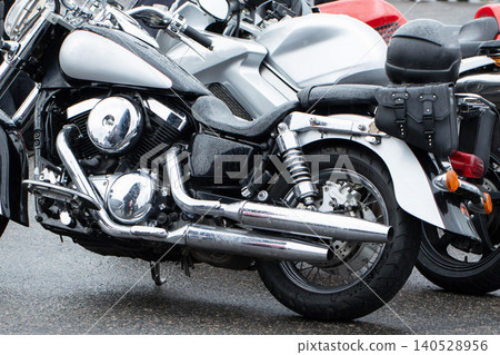 Close-up detail of modern cruiser motorcycle parked on wet asphalt surface on rainy day, chrome accents reflecting light, alongside other motorcycles and a black helmet, showcasing biker culture 140528956