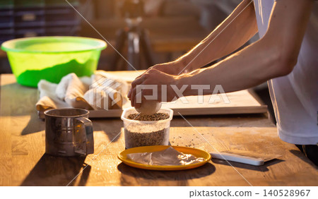 Skilled baker hands meticulously prepare dough, rolling ball in mixture of seeds on rustic wooden table. Golden light highlights culinary craftsmanship during baking process. Skilled baker hands meticulously prepare dough, rolling ball in mixture of seeds on rustic wooden table. Golden light highlights culinary craftsmanship during baking process. 140528967