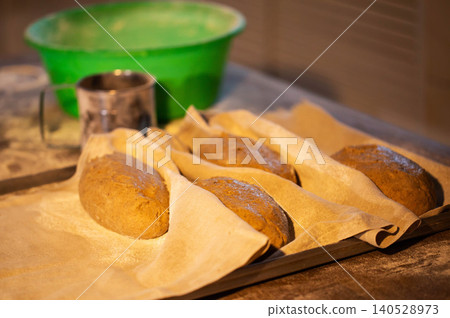 Freshly kneaded artisan whole grain bread loaves prepared for baking on parchment paper lined tray in bakery kitchen with green bowl background Freshly kneaded artisan whole grain bread loaves prepared for baking on parchment paper lined tray in bakery kitchen with green bowl background 140528973