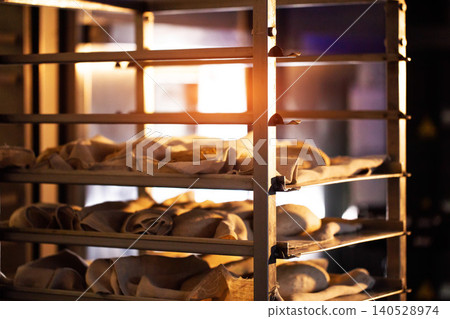 Unbaked bread dough loaves proofing on multiple shelves of bakery rack inside bread factory producing artisanal baked goods by hand with warm light Unbaked bread dough loaves proofing on multiple shelves of bakery rack inside bread factory producing artisanal baked goods by hand with warm light 140528974