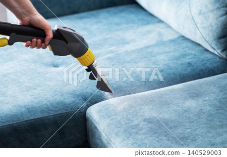Hand operates professional cleaning tool spraying solution onto blue fabric sofa, performing deep cleaning for upholstery sanitization and freshness. Hand operates professional cleaning tool spraying solution onto blue fabric sofa, performing deep cleaning for upholstery sanitization and freshness. 140529003