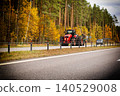 Red agricultural tractor with attached trailer driving on asphalt road, green roadside barrier, vibrant autumn trees and evergreen forest lining background, black SUV passing opposite lane, rural 140529008