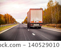White semi-truck trailer driving on highway during autumn sunset. Road transport of cargo vehicle on open road with fall season trees under cloudy sky. 140529010