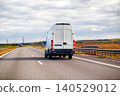 White cargo van driving on highway under cloudy sky during day, transport vehicle moving on asphalt road with fields and trees in background 140529012