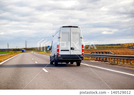 White cargo van driving on highway under cloudy sky during day, transport vehicle moving on asphalt road with fields and trees in background White cargo van driving on highway under cloudy sky during day, transport vehicle moving on asphalt road with fields and trees in background 140529012