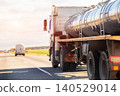 Rear view of large tanker truck transporting liquid cargo on highway during golden hour. Sunset or sunrise light illuminates metal tank, long shadows stretch across asphalt road, vehicle moving away 140529014