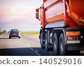 Red heavy dump truck traveling on highway road under bright sky at sunset or sunrise with car in distance 140529016