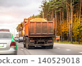 Rear view of orange dump truck carrying sand on highway with other vehicles in traffic, autumn forest lining side of road under overcast sky, transportation of construction materials. 140529018