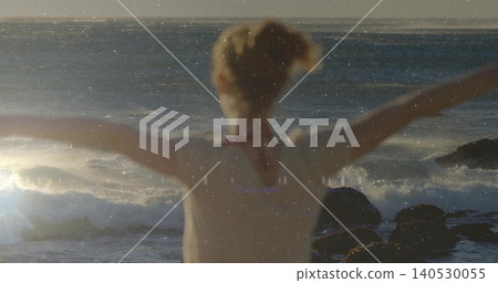 Standing woman from back stretching arms on rocky shore, facing sunlit waves, in red strap top Standing woman from back stretching arms on rocky shore, facing sunlit waves, in red strap top 140530055