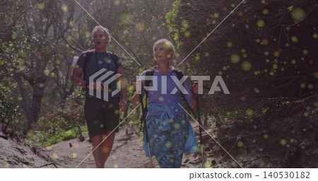 Walking senior couple wearing navy shirt blue top on sandy trail, carrying daypacks camera poles Walking senior couple wearing navy shirt blue top on sandy trail, carrying daypacks camera poles 140530182
