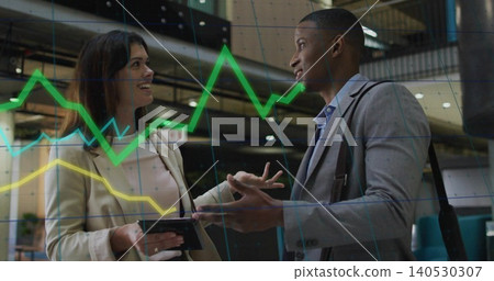 Discussing pair in business attire with tablet and shoulder bag under faint chart overlay in atrium Discussing pair in business attire with tablet and shoulder bag under faint chart overlay in atrium 140530307