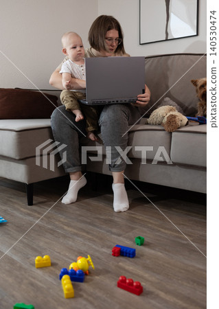 Parent on couch holding baby while using laptop with toys on floor 140530474