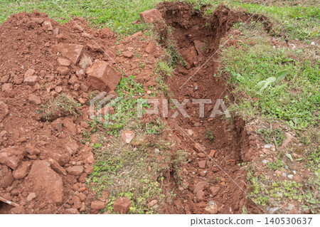 A high angle view of an open earthworks trench split junction dug into dark brown soil and green lawn grass for multi-line residential utility layout. Underground infrastructure pipe intersection 140530637