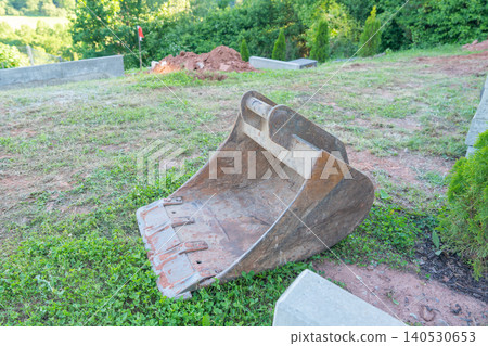 A heavy steel excavator bucket detached and sitting on green grass at a residential construction site with earthworks in background. Industrial digger attachment machine component placed on ground A heavy steel excavator bucket detached and sitting on green grass at a residential construction site with earthworks in background. Industrial digger attachment machine component placed on ground 140530653