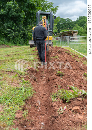 Vertical full shot of a construction worker standing inside a freshly dug dirt trench, manually aligning a flexible black service utility pipe. Residential backyard landscaping and drainage system Vertical full shot of a construction worker standing inside a freshly dug dirt trench, manually aligning a flexible black service utility pipe. Residential backyard landscaping and drainage system 140530665