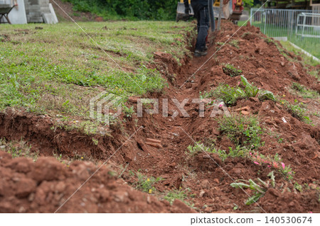 Close-up of a long narrow trench dug into brown earth on a green lawn hillside for pipeline installation or drainage foundation. Excavated ground soil groove layout prepared during backyard 140530674