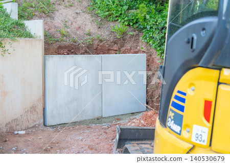 Prefabricated concrete retaining wall panels installed into the ground next to a yellow mini excavator at a residential site. Modern backyard landscaping showing concrete block support sections for 140530679