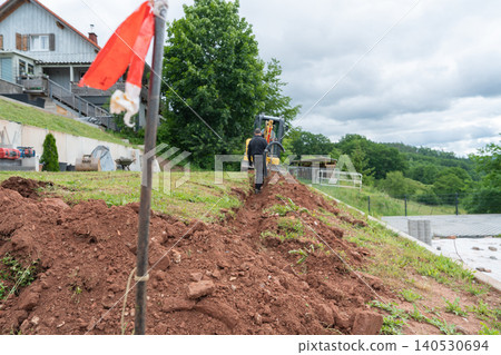 A long open construction trench stretch on a grassy hill profile with a red safety boundary flag marking the excavation zone. Residential garden renovation project featuring terrain modeling A long open construction trench stretch on a grassy hill profile with a red safety boundary flag marking the excavation zone. Residential garden renovation project featuring terrain modeling 140530694