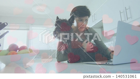 Holding terrier, woman in longsleeve top typing on laptop at kitchen counter, pink heart overlay Holding terrier, woman in longsleeve top typing on laptop at kitchen counter, pink heart overlay 140531061