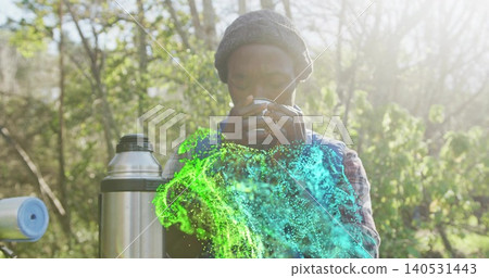 Sipping man in beanie and vest standing in sunlit woods, holding cup near green cyan mist Sipping man in beanie and vest standing in sunlit woods, holding cup near green cyan mist 140531443