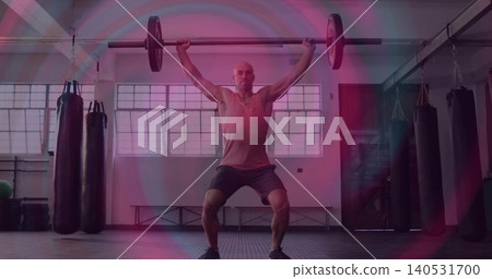 Mid-adult male in tank top, shorts, shoes holding loaded barbell overhead in gym with heavy bags Mid-adult male in tank top, shorts, shoes holding loaded barbell overhead in gym with heavy bags 140531700