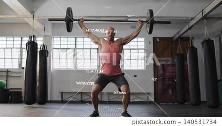 Lifting man pressing Olympic barbell overhead in gym with hanging bags, wearing red tank top 140531734