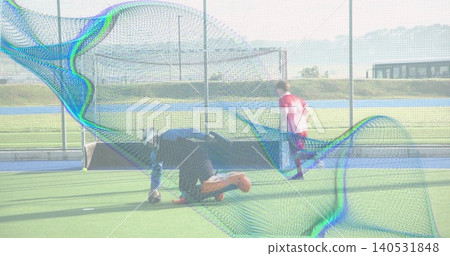 Kneeling goalkeeper in helmet and glove reaching for ball on synthetic turf pink-clad player racing Kneeling goalkeeper in helmet and glove reaching for ball on synthetic turf pink-clad player racing 140531848