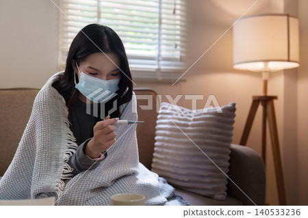 Asian woman checking temperature with mask on sofa at home for fever Asian woman checking temperature with mask on sofa at home for fever 140533236