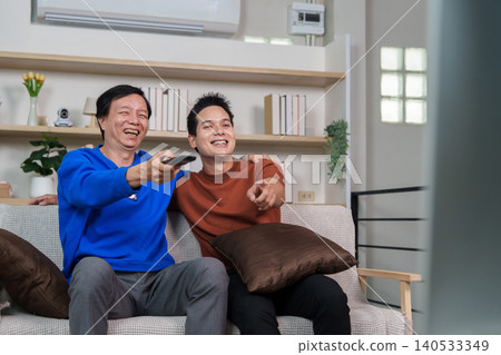 East Asian father and son laughing watching TV in living room East Asian father and son laughing watching TV in living room 140533349