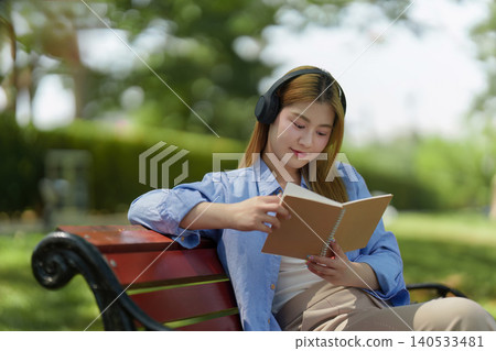 Asian woman reading book with headphones on park bench, self-care Asian woman reading book with headphones on park bench, self-care 140533481