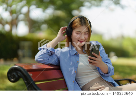 Asian woman in 20s listening to music on headphones in park Asian woman in 20s listening to music on headphones in park 140533640