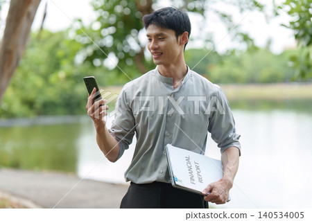 East Asian man reviewing financial report and smartphone outdoors East Asian man reviewing financial report and smartphone outdoors 140534005