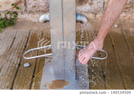Barefoot person rinsing sandy foot at public beach shower near Mediterranean coast 140534509