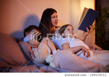 Mother reading bedtime story to two children on sofa in warm evening light. Mother reading bedtime story to two children on sofa in warm evening light. 140534511
