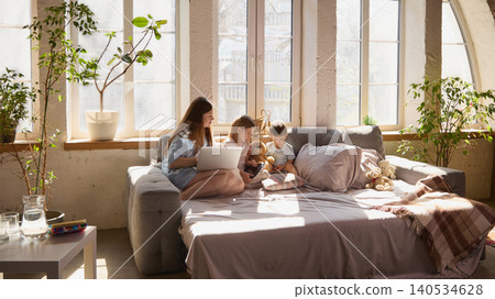 Wide shot of mother and two children using laptop and tablet on sofa in bright living room. Wide shot of mother and two children using laptop and tablet on sofa in bright living room. 140534628