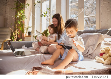 Little boy focused on smartphone while mother and sister use laptop on sofa in sunny living room. Little boy focused on smartphone while mother and sister use laptop on sofa in sunny living room. 140534641