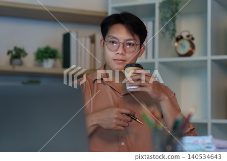 East Asian man focused on online meeting with laptop and coffee cup at night East Asian man focused on online meeting with laptop and coffee cup at night 140534643