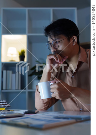 Thoughtful Asian man contemplating with coffee mug working late at home office Thoughtful Asian man contemplating with coffee mug working late at home office 140534842